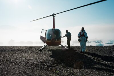 A bride disembarks from a helicopter, silhouetted in the fog coming off the glacier lagoon. they are coming from exchanging vows during their Alaska Glacier Elopement wedding.  A guide closes the door behind her.  She looks regal in the  frame.