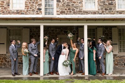 The bride and groom share a kiss under a covered porch as their wedding party cheers around them. Bridesmaids in shades of green lift their bouquets, while groomsmen in sleek gray suits raise their fists in celebration. The rustic stone exterior of the Pennsylvania venue serves as a timeless setting for this moment of pure joy