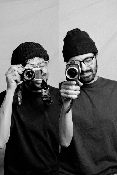 Black-and-white split portrait of two photographers with vintage cameras, wearing beanies and glasses. One holds a Canon film camera, the other a classic video camera, capturing timeless moments with an analog touch.