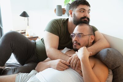 This image shows two male presenting partners lying together in bed. One partner is staring ahead, past the camera, with a serious expression. The other partner, with a beard and a nose ring, wraps their arms around the partner, lying above them. They look away in the other direction.