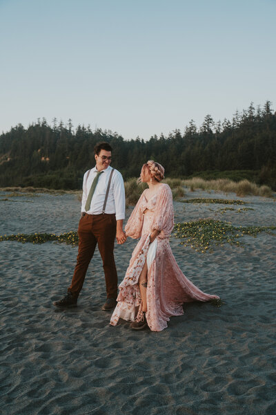 Bride and groom hold hands and walk down beach looking at each other after eloping in california by california elopement photographer kasey mantiply