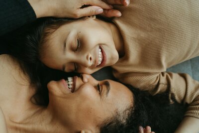Mother and daughter lay on the ground and smile while facing one another.