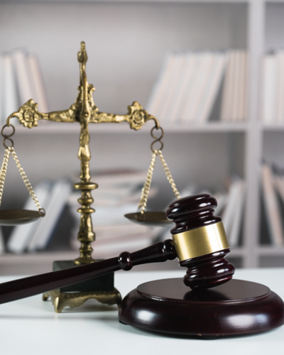 Close-up of a wooden judge's gavel resting beside brass balance scales, with law books blurred in the background.