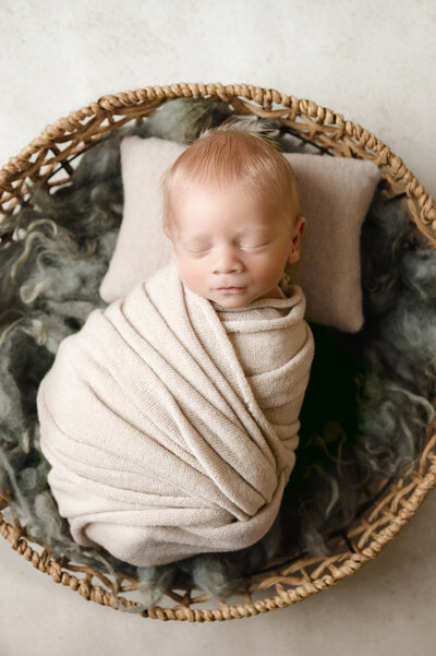 Newborn boy is wrapped in a tan blanket and laying on a pillow in a round wicker basket.