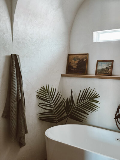 Serene, minimalist bathroom with arched plaster walls, a freestanding soaking tub, vintage art, and palm leaves, designed to evoke calm and natural wellness.


