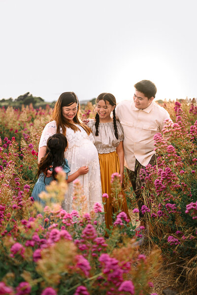 family of 4 all looking at a pregnant mama in a white maternity dress in a field of purple wildflowers