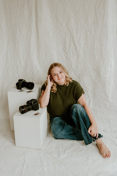 A woman with blonde hair sits on a wooden floor against a plain wall, wearing a green jacket and holding a camera. She smiles, creating a relaxed, joyful mood.