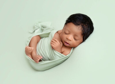 Baby Boy is wrapped in a white knit swaddle for a newborn photoshoot. Baby's hands are peeking out of the top of the swaddle holding a petite white teddy bear. Baby is sleeping.