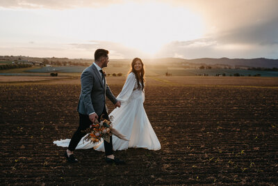 bride and groom walking in a field as the sun sets