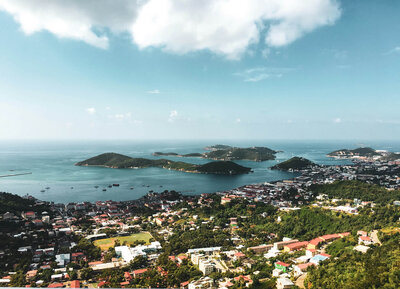 Discover an aerial view of a coastal city at epicsandstravel.com/about, where lush greenery and scattered rooftops contrast with the azure ocean. Small, verdant islands lie on the horizon under a partly cloudy sky, inviting exploration and adventure.
