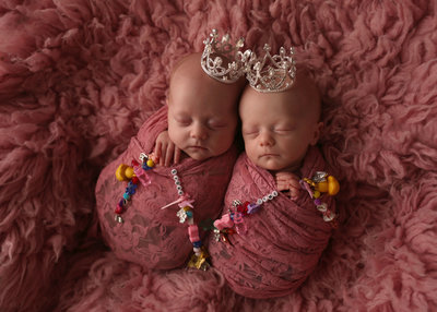 twin newborns both wrapped in rose pink lace with tiaras  holding beaded charms laying on pink fur rug