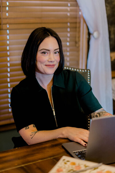 A person with shoulder-length dark hair, wearing a dark zip-up top, sits at a wooden desk using a laptop. They are smiling and have tattoos on their left arm. The background features closed wooden blinds and a partially visible curtain.