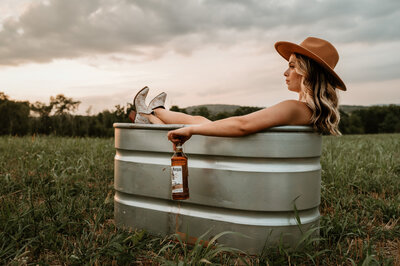 Woman sits in a water trough with cowgirl boots and a bottle of whiskey. 