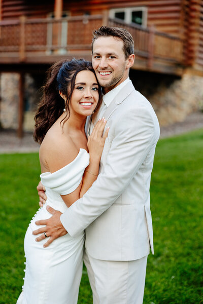 A smiling couple in formal attire stands on a grassy lawn, captured beautifully by a Minnesota photographer. The woman in a white off-shoulder gown and the man in a light suit embrace before a wooden building, creating an enchanting Brainerd Lakes photography moment with a slightly blurred background.