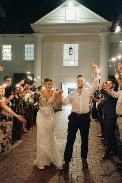Bride and groom celebrating during a sparkler exit at Adams Estate in Florida, walking hand-in-hand through cheering guests at night — bride in a beaded gown and groom with rolled sleeves, captured in a fun and candid wedding moment.