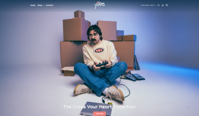 A man with dark hair, a mustache and glasses sitting on the floor playing an old video game with stacked boxes in the background.