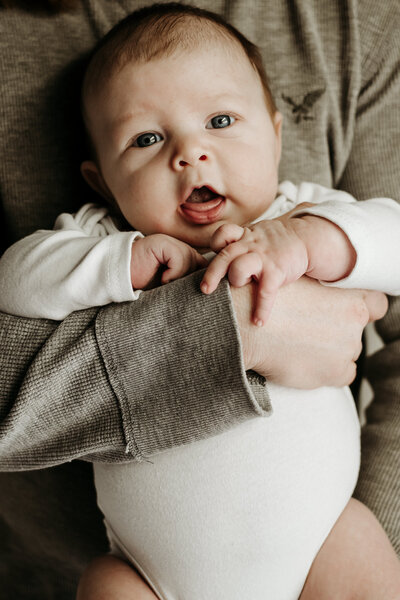 Three month old baby boy wearing white while dad is holding him in moody studio.