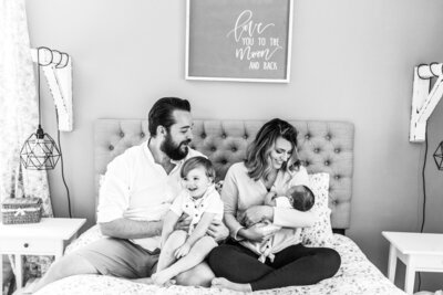 A black and white photo of Reg and Kala holding Elliott and their newborn son Oliver at home on their bed.