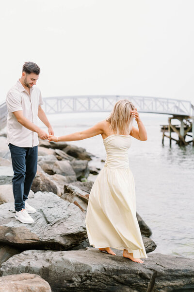 Newport Rhode Island Engagement Photo Photographer | A woman in a white dress and a man in a casual shirt hold hands on rocky waterfront. Background features a bridge.