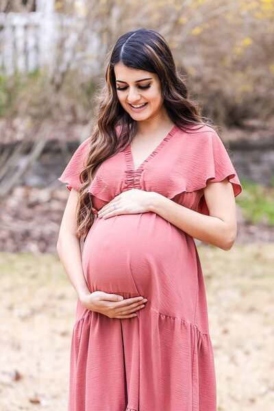 New mom in rose colored maternity dress holding her belly smiling as she awaits the arrival of her new little one