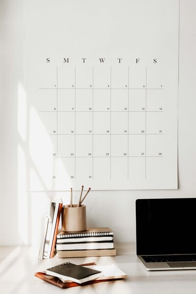 white wall with large white calendar, black  day and month letters, light oak desk, open macbook pro, stacks of books, jars of pencils, notepads