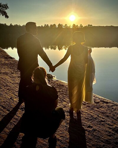 Beautifil sunset on a lake with a brideand groom holding hands and looking at each other while a woman is crouched getting a shot of the sun