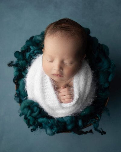 Newborn boy sleeping, wearing white wrap, on dark teal background, with hands together at chest