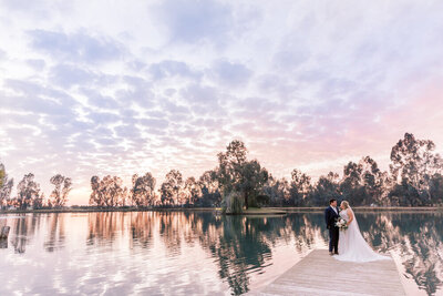 bride and groom standing on dock by lake at sunset