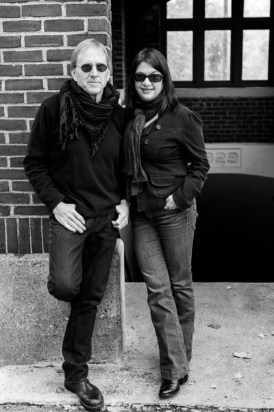 Black and white branding photo of Open Book Music duo Rick and Michele Gedney leaning against brick wall in downtown Cold Spring, NY near train station.