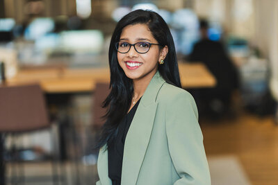 Professional corporate headshot of woman with glasses and long dark hair, wearing green blazer in office setting