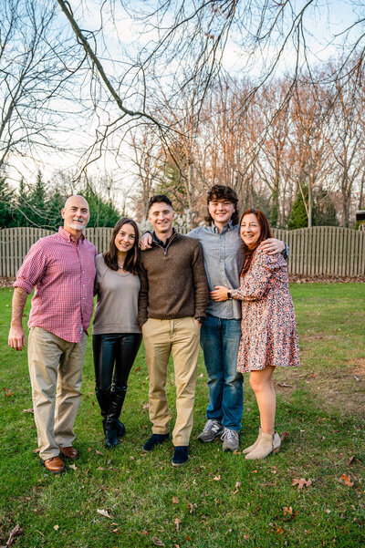 Family of five, including their 3 young adult children, standing in their backyard, smiling while hugging next to one another.