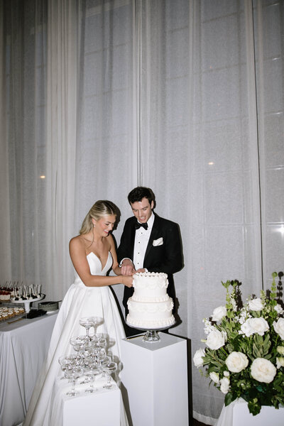 Bride and groom cutting a tiered wedding cake with smiles. Bride in white gown; groom in black tuxedo. Elegant setup with flowers and glasses nearby.