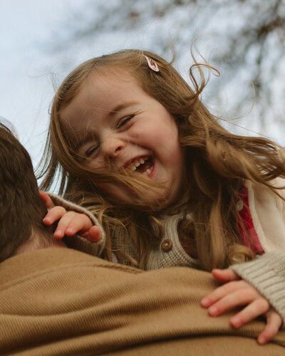 little girl laughs as her father tosses her over his shoulder on a family walk in the woods