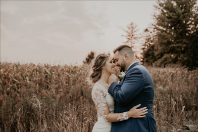 Bride and groom are standing for a wedding day portrait in a field in London, ON. The bride has her arms wrapped around her groom's waist. The groom is tilting the bride's chin up to give her a kiss. Both are smiling at each other with their eyes closed. Captured by top London-ON wedding photographer Ashlee Ellison.