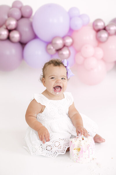 Baby girl sitting on a white backdrop in front of purple and pink balloons smiling as she smashes into her first birthday cake