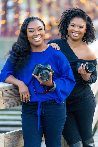 Two African American women pose with their cameras in hand on a walkway.