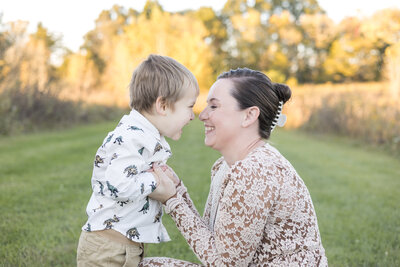 mom knelt down to young son, looking at each other laughing.  Son has sandy short blonde hair, and mom's dark hair is pulled back, they are holding each other's hands