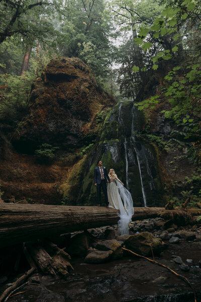 Bride and groom walk on large log in front of waterfall in Umpqua National Forest