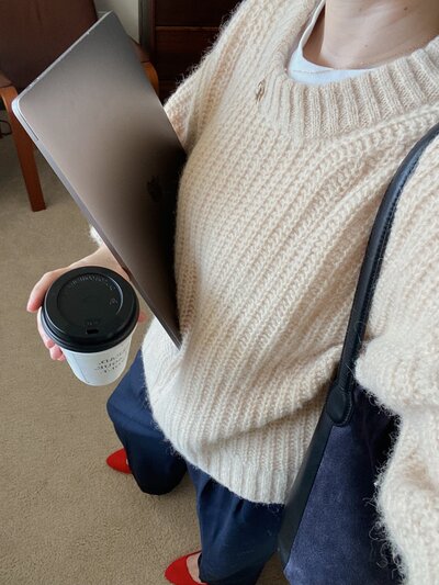 Photo taken from a selfie angle of a woman wearing a sweater and holding a to-go coffee and a laptop