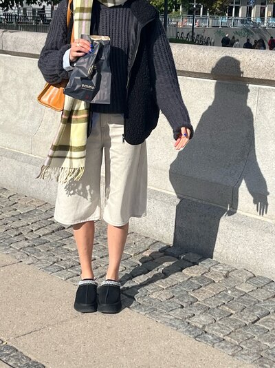 A person stands by a stone wall, wearing a cozy black sweater, white culottes, and slippers, holding a shopping bag. A plaid scarf and bright sunlight cast shadows.