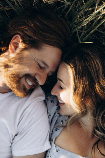 A close-up image from a photoshoot of a smiling man and woman lying on grass, their heads touching as they share a joyful moment, highlighting their emotional connection and happiness.