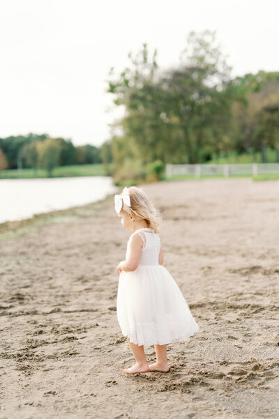 Little girl in a white dress standing on the beach, looking out at the water.