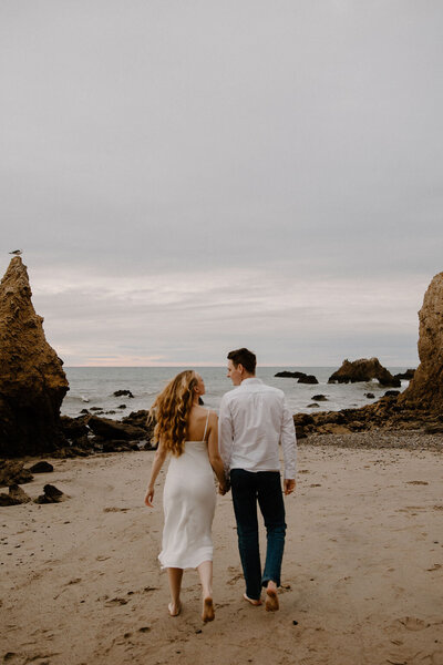 Engagement photography session captured by Morgan Ashley Lynn Photography in Milwaukee, WI on the beach of Lake Michigan. The couple are walking toward the water on the sand holding hands and looking at each other. The bride to be has long blond curly hair and is wearing a white dress and future groom is wearing jeans and a white button down