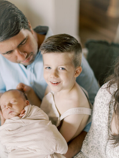 Brunette boy in cream overalls holds newborn baby brother in cream swaddle while sitting in brunette dad's lap at their Little Rock home taken by Bailey Feeler Photography