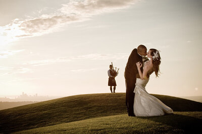 Bride and groom kiss on hill in front of bag pipe player