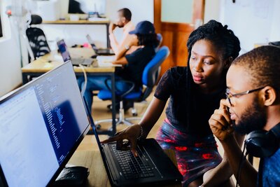 2 people working at a computer in a room with others working at different tables.