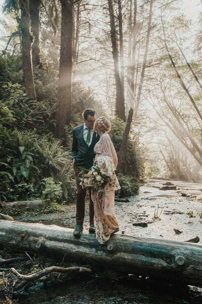 Bride and groom turned towards each other looking at each other standing on a log in a canyon surrounded by trees and light rays by Big Sur elopement photographer Kasey Mantiply