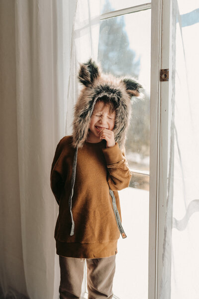 Child makes silly face wearing a wolf hat standing beside a window
