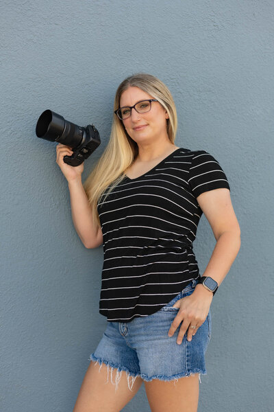 Megan Bowers, a professional senior photographer, poses confidently against a textured blue wall while holding her camera. Her relaxed yet bold stance reflects her artistic and modern approach to capturing authentic senior portraits. Megan Bowers Photography specializes in expressive and timeless senior photography.