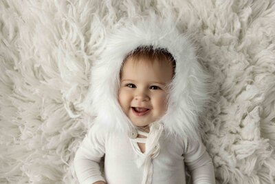 Baby photoshoot from London, Ontario lying on cream fluffy rug wearing a white knit onesie and faux-fur trimmed bonnet. Baby has a big smile and starting right at the camera